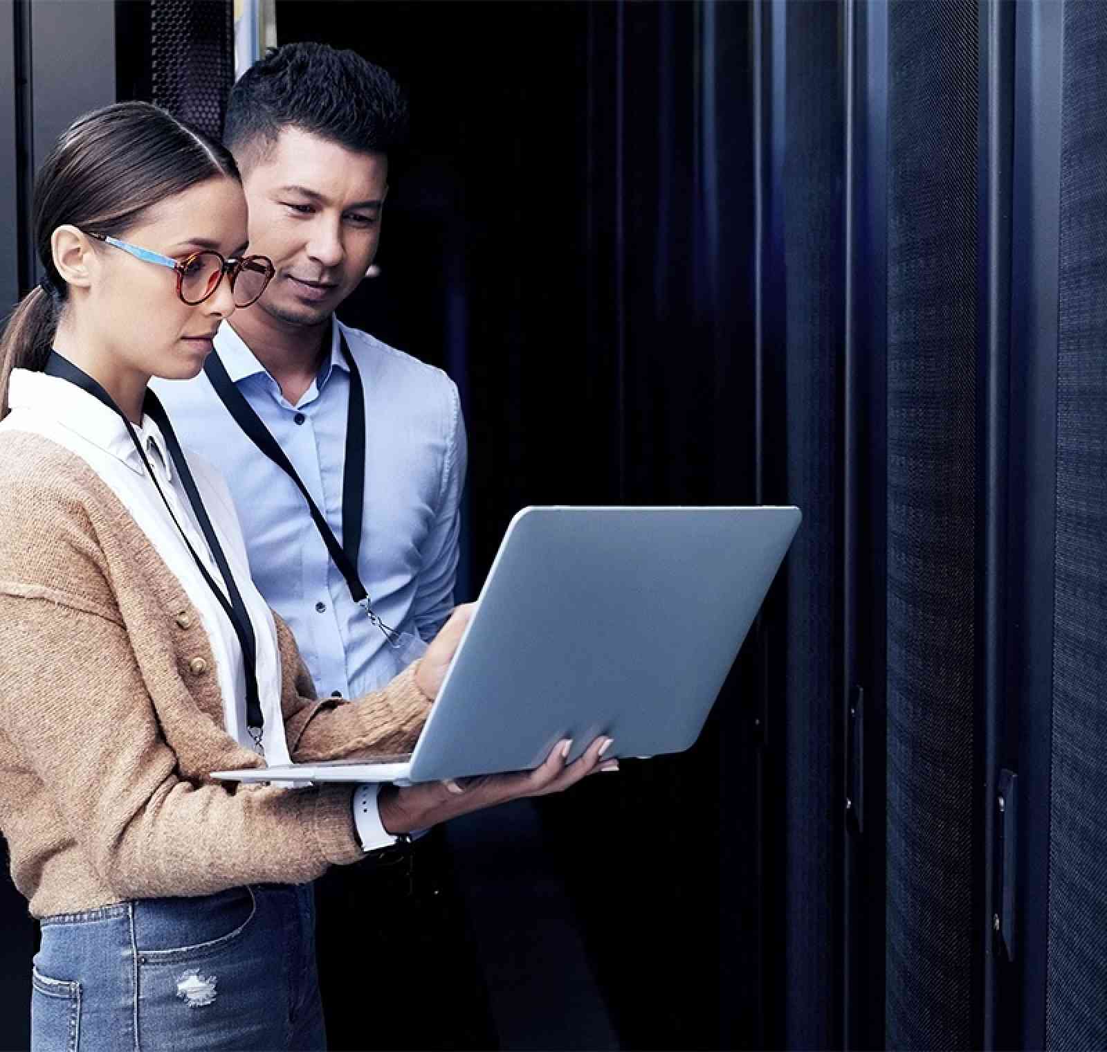 An IT professional stands confidently in a modern office surrounded by technology, reviewing data on a tablet while computer monitors display system code and network analytics in the background.